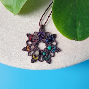 May include: A brown macrame necklace with a seven-chakra crystal pendant. The pendant is a circular design with seven different colored stones in the center.