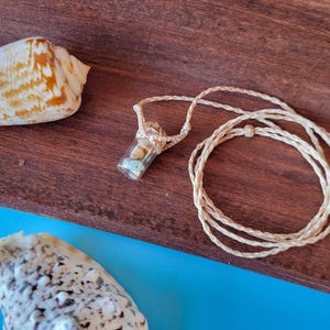 May include: A necklace with a small glass vial filled with seashells, hanging from a natural fiber cord. The necklace is displayed on a wooden surface with seashells.