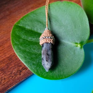 May include: A handcrafted pendant featuring a faceted, gray and black crystal. The crystal is adorned with a tan macrame cap, accented with silver-colored beads, and suspended from a matching cord. The pendant rests on a large green leaf.
