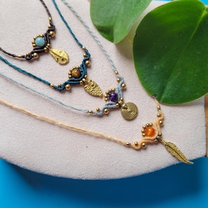 Könnte beinhalten: Drei übereinanderliegende Halsketten mit verschiedenen farbigen Steinen und Anhängern. Die oberste Halskette hat einen hellblauen Stein und einen Federnanhänger. Die mittlere Halskette hat einen Tigerauge-Stein und einen Blattanhänger. Die unterste Halskette hat einen violetten Stein, einen Spiral-Anhänger und einen Federnanhänger.