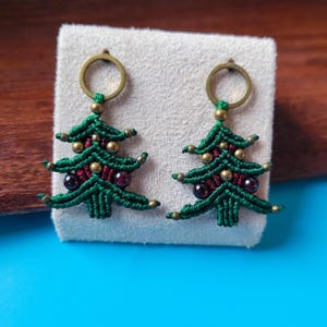 May include: A pair of handmade macrame Christmas tree earrings. The earrings are green with red and gold accents, and feature a brass ring at the top for hanging. The earrings are displayed on a white surface against a blue background.