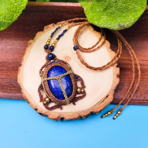 May include: A handcrafted necklace featuring a large, oval, deep blue lapis lazuli stone. The stone is encased in a macrame design with brown and gold accents. The necklace hangs on a brown cord, presented on a wooden slice.