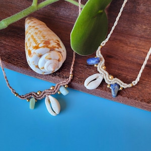 May include: Two handmade macrame necklaces with shell and gemstone accents. The necklace on the left has a brown cord with green stones and a white shell. The necklace on the right has a white cord with blue stones and a white shell.