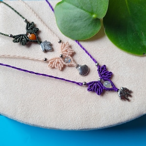 May include: Three necklaces with different colored cords and charms. The top necklace has a green cord with a small orange stone and leaf charm. The middle necklace has a cream cord with a seashell charm and a white stone. The bottom necklace has a purple cord with a white stone and a small silver charm.