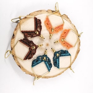 May include: Six pairs of handmade earrings with geometric designs. The earrings are made with brown, orange, and teal thread and gold beads. The earrings are hanging from gold hoops.