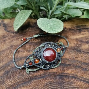 Puede incluir: Un brazalete de macramé gris con una gran piedra de cornalina roja y pequeños detalles de cornalina. El brazalete está atado con un cordón gris y tiene cuentas doradas.