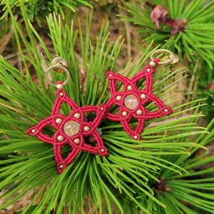 May include: A pair of handmade red macrame star earrings with gold-colored accents and a central clear stone. The earrings are displayed on a bed of green pine needles, showcasing the intricate knotting and design.