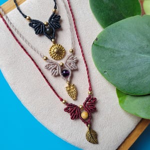 Puede incluir: Tres collares superpuestos con cordones y dijes de diferentes colores. El collar superior tiene un cordón negro con un dije de búho negro y dorado. El collar del medio tiene un cordón beige con un dije circular dorado y una piedra morada. El collar inferior tiene un cordón marrón con un dije de búho dorado y una piedra marrón.