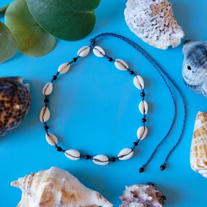 May include: A white shell necklace with black beads on a blue background. The necklace is made of cowrie shells and is tied with a blue cord.