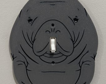 Può includere: Una copertura per interruttore della luce a forma di lamantino grigio con un interruttore bianco. La copertura ha contorni neri ed è montata su una parete grigio chiaro. Il design del lamantino include occhi, naso e bocca sorridente.
