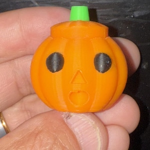 May include: A 3D-printed orange pumpkin with black eyes and a surprised expression. The pumpkin has a green stem and is a festive decoration for Halloween. The pumpkin is held in a hand.