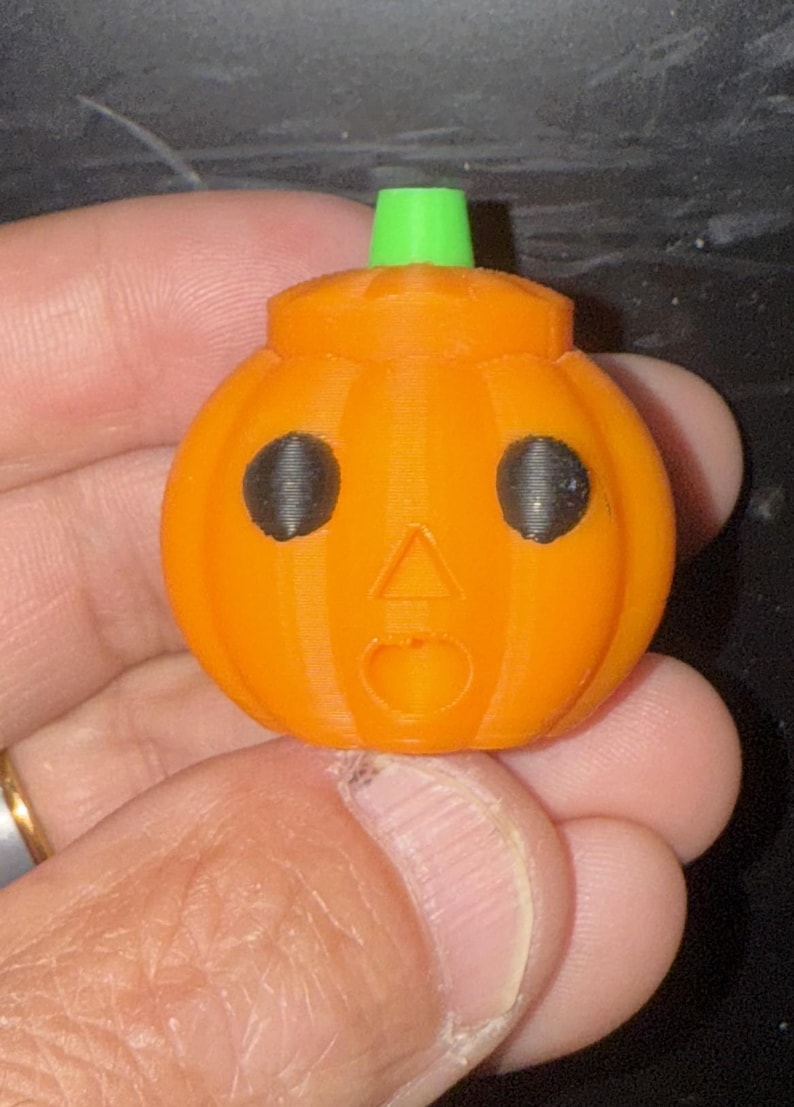 May include: A 3D-printed orange pumpkin with black eyes and a surprised expression. The pumpkin has a green stem and is a festive decoration for Halloween. The pumpkin is held in a hand.