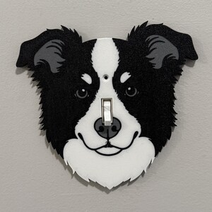 May include: A decorative light switch cover shaped like a dog's face. The dog has black and white fur, with a white muzzle and a black nose. The switch is a standard toggle style.