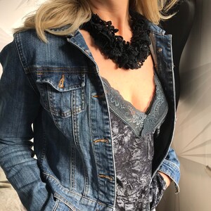 May include: A woman wearing a denim jacket over a gray velvet top with lace trim. She is wearing a black lace choker necklace.