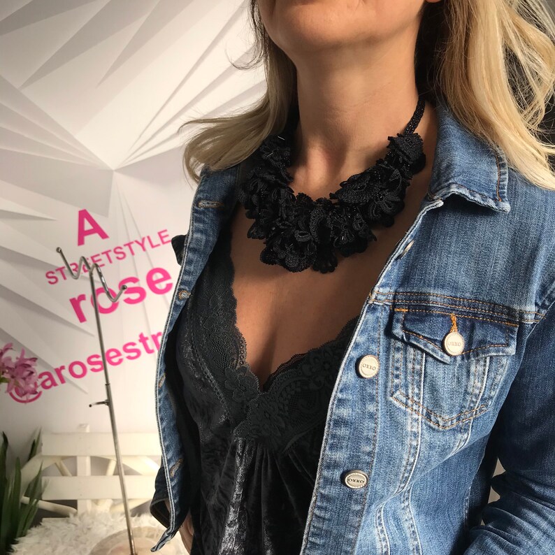 May include: A woman wearing a black lace top and a denim jacket with a large black statement necklace. The necklace is made of many layers of fabric and has a ruffled appearance.