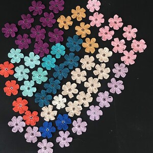 May include: Assortment of colourful crocheted flower embellishments in various shades of purple, blue, green, pink, and orange. The flowers are arranged in a circular pattern on a black background.