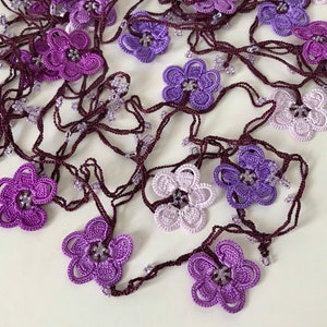 May include: A purple and lavender crocheted flower necklace with a brown cord. The necklace is made of delicate crocheted flowers in various shades of purple and lavender.
