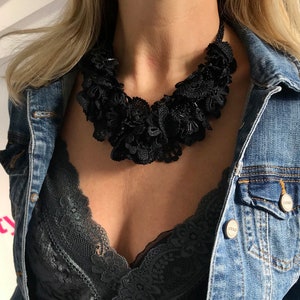 May include: A black lace choker necklace with a layered, ruffled design. The necklace is worn by a person with blonde hair and a denim jacket.