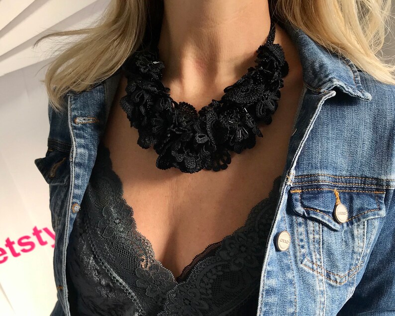 May include: A black lace choker necklace with a layered, ruffled design. The necklace is worn by a person with blonde hair and a denim jacket.