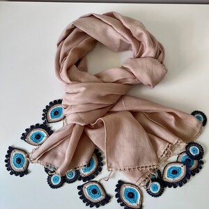 May include: A blush pink scarf with a crocheted trim featuring blue evil eye charms.