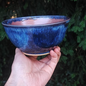 May include: A ceramic bowl featuring a deep blue glaze with lighter blue and purple streaks. The interior of the bowl reveals a warm, earthy terracotta tone. The bowl has a smooth, glossy finish and a rounded shape with a visible unglazed base.