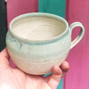 May include: A round ceramic mug with a speckled off-white base and a light blue glaze dripping from the rim. The mug features a curved handle on the right side and a wide, open top. The surface has a textured, matte appearance with visible clay grain.