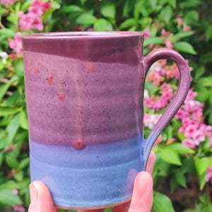 May include: A handcrafted ceramic mug with a colour gradient. The top is a deep burgundy, fading into a medium purple, and then a light blue at the base. It has a curved handle and is set against a backdrop of green foliage and pink flowers.