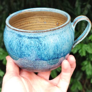 May include: A round ceramic mug featuring a speckled blue glaze that transitions from a deep navy to a lighter sky blue. The interior of the mug is a smooth, unglazed tan colour with visible horizontal ridges along the inner walls and a single handle.