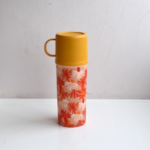 May include: A cylindrical plastic travel mug with a solid mustard yellow lid featuring a side handle. The body of the mug is decorated with a repeating pattern of stylized red and white daisy-like flowers against a light peach background.