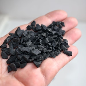 May include: A close-up of a hand holding a collection of small, dark grey decorative stones. The stones are irregularly shaped with sharp edges and a matte finish. The background is plain white.