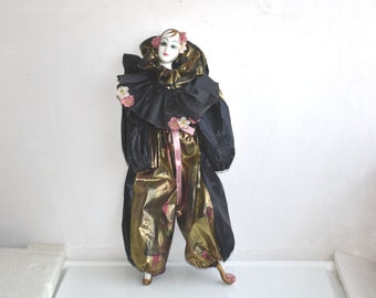 May include: A porcelain clown doll with a black and gold costume. The doll has a white face with painted features and pink flowers in its hair. The costume has a large black collar and gold pants with pink accents.