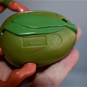 May include: Close-up of a green and orange action figure. The figure's back is olive green with a green leaf-like detail. The text "Made in China" and "NG1606A" are visible. The figure's arms and legs are orange.