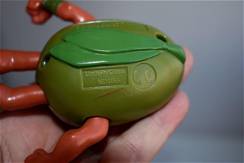 May include: Close-up of a green and orange action figure. The figure's back is olive green with a green leaf-like detail. The text "Made in China" and "NG1606A" are visible. The figure's arms and legs are orange.