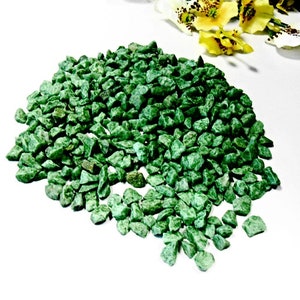 Puede incluir: Un montón de pequeñas piedras verdes de forma irregular. Las piedras son de un tono verde vibrante y parecen estar trituradas o rotas. Las piedras están esparcidas sobre una superficie blanca, con flores amarillas y blancas en el fondo.