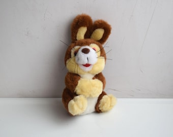 May include: A soft toy rabbit with brown and beige fur, a white face, and a red mouth. The rabbit has long ears and black whiskers. The toy is sitting upright with its paws crossed. A classic children's toy.