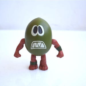 May include: A green, egg-shaped figurine with a white-outlined face featuring black eyes and a jagged mouth. The figurine has red arms and legs with green bands. A unique, whimsical collectible.