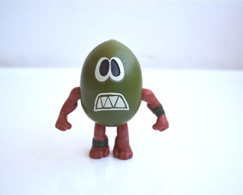 May include: A green, egg-shaped figurine with a white-outlined face featuring black eyes and a jagged mouth. The figurine has red arms and legs with green bands. A unique, whimsical collectible.