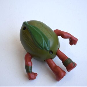 May include: A vintage olive-shaped toy figure with red arms and legs. The figure has a green leaf detail on top and green bands around the ankles. The toy is on a white surface.
