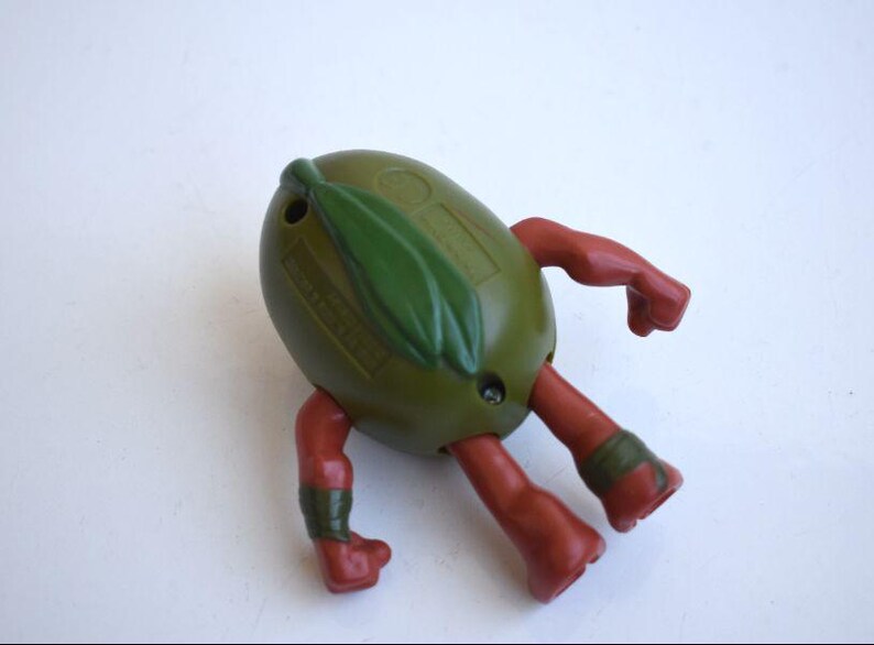 May include: A vintage olive-shaped toy figure with red arms and legs. The figure has a green leaf detail on top and green bands around the ankles. The toy is on a white surface.