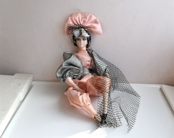 May include: A porcelain doll with black hair and a pink and silver dress. The doll is wearing a pink hat with a black veil and a black net skirt.