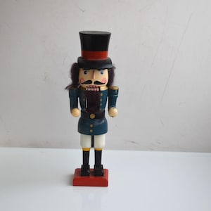 Puede incluir: Figura de cascanueces de madera con forma de soldado. La figura tiene un sombrero de copa negro con una banda roja, una chaqueta azul y pantalones blancos. Se apoya sobre una base roja y tiene una cara detallada con bigote.