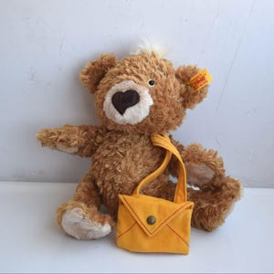 May include: A brown teddy bear with a heart-shaped nose and a small yellow satchel. The bear has a cream-colored muzzle and a tuft of white fur on its head. The satchel has a button closure and a yellow strap.