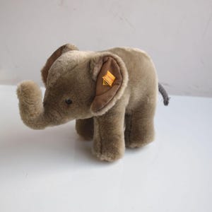 Può includere: Un elefante di peluche nei toni del marrone e del beige. L'elefante ha grandi orecchie, una proboscide e una piccola coda. Un'etichetta gialla è attaccata a un orecchio. Il giocattolo è realizzato in materiale morbido e soffice.