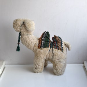 May include: A white plush camel toy with a colourful patterned saddle. The camel has a green tassel hanging from its neck.