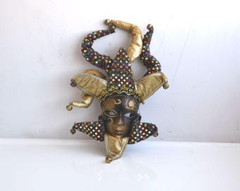 May include: A gold and black jester mask with colorful sequins and bells. The mask has a pointed hat with two long, curled horns.