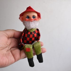 Pode incluir: Uma boneca vintage com um chapéu vermelho, barba branca e camisa xadrez vermelha e preta. A boneca tem olhos azuis, bochechas rosadas e pernas verdes e pretas. A boneca está sentada.