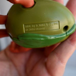May include: A green and orange toy, possibly a character from a children's show, held in a hand. The toy has a rounded shape with a green shell-like design and orange limbs. The text "Mfd. by b. little & co., Inc. &copy; 2016 Disney" is visible.