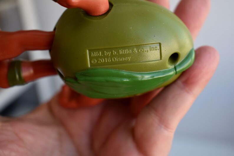 May include: A green and orange toy, possibly a character from a children's show, held in a hand. The toy has a rounded shape with a green shell-like design and orange limbs. The text "Mfd. by b. little & co., Inc. &copy; 2016 Disney" is visible.