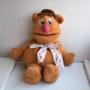 May include: A plush toy of Fozzie Bear, a Muppet character, with a brown hat, light brown fur, and a pink nose. He wears a white bow tie with pink polka dots. The toy is sitting down.