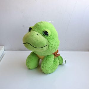 Könnte beinhalten: Ein grünes Plüschtier in Form einer Schildkröte mit einem lächelnden Gesicht, schwarzen Augen und einem braun-weißen Schalenmuster. Das Stofftier besteht aus weichem Material und ist für Kinder konzipiert.